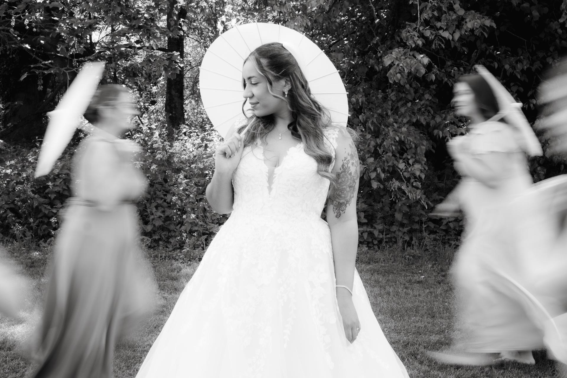 black and white portrait of a bride with a white umbrella, bridesmaids walking blurred in the background, Suffolk Weddings, Heffs Photography, 